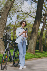 Full length young smiling fun woman in jeans clothes stand near bicycle bike on sidewalk in city spring park outdoors use mobile cell phone chat online. People urban healthy lifestyle cycling concept.