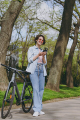 Fototapeta premium Full length young smiling fun woman in jeans clothes stand near bicycle bike on sidewalk in city spring park outdoors use mobile cell phone chat online. People urban healthy lifestyle cycling concept.