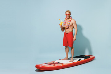 Full length young sexy man 20s in red shorts swimsuit hawaii lei glasses stand on sup board hold...