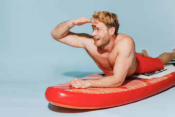 Young sexy strong sporty man in red shorts swimsuit lying on sup board paddle hold hand at forehead...