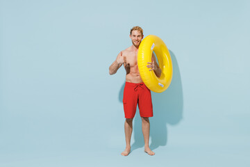 Full length young sexy sporty man in red shorts swimsuit relax near hotel pool hold inflatable ring show thumb up gesture isolated on pastel blue background. Summer vacation sea rest sun tan concept