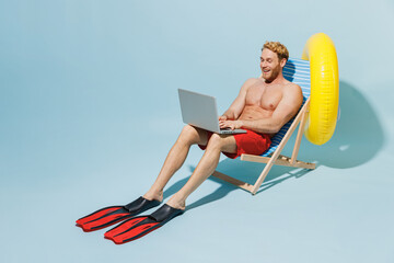 Full length young freelancer man in red shorts swimsuit sit in deckchair near inflatable ring using laprop pc computer work isolated on pastel blue background Summer vacation sea rest sun tan concept