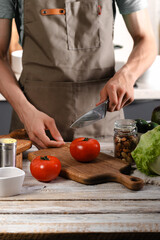 A young chef in a brown apron uses a kitchen knife to cut vegetables for a delicious salad. Cooking delicious and healthy food in domestic kitchen. Freshnes vegetarian food. Vertical shot. Copy space