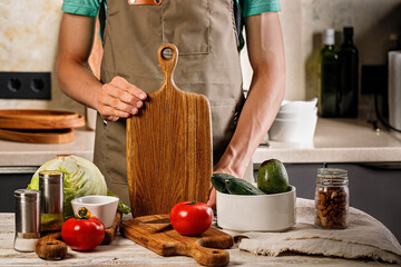 A young chef in a brown apron holds a wooden cutting board. Vegetables for a vegetarian salad are on the table. Cooking delicious and healthy food in your home kitchen.
