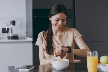 Fotobehang Young smiling happy punctual calm housewife woman 20s wear beige t-shirt eat breakfast in morning look at smart watch check time cooking food in light kitchen at home Healthy diet lifestyle concept. © ViDi Studio