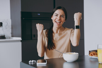 Young happy successful overjoyed housewife woman 20s in beige tshirt eat breakfast in morning look camera use smart watch clench fist cook food in light kitchen at home Healthy diet lifestyle concept