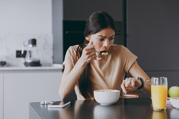 Fototapeta na wymiar Young sad displeased disappointedhousewife woman 20s in t-shirt eat breakfast in morning look at smart watch check time hurry rush cooking food in light kitchen at home Healthy diet lifestyle concept.