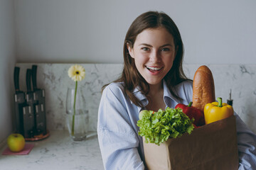 Young surprised vegeterian caucasian housewife woman 20s in casual clothes blue shirt hold paper bag with seasonal vegetable cooking food in light kitchen at home alone Healthy diet lifestyle concept.