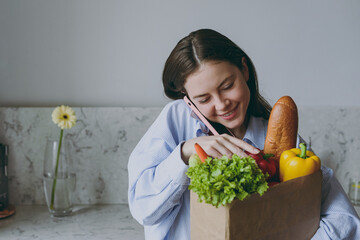 Young smiling vegeterian housewife woman 20s in casual clothes blue shirt hold bag with vegetables talk by mobile cell phone cooking food in light kitchen at home alone Healthy diet lifestyle concept.