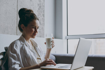 Young side view smart freelancer woman in white clothes sit on grey sofa near window indoor apartment work online laptop pc computer drink milk Rest on weekends leisure quarantine stay home concept.