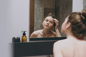 Back rear view nice half naked topless caucasian young woman 20s touching forehead reflected in mirror in bathroom doing morning routine. Skin care healthcare cosmetic procedures lifestyle concept