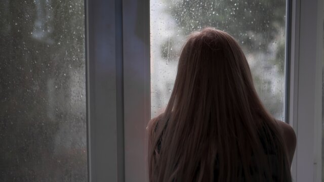 An Unrecognizable, Bored, Lonely Girl Sits At Home Due To Bad Rainy Weather And Looks Out The Window At The Falling Raindrops. Wait For Good Weather. Back View.