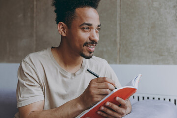 Dreamful creative happy young student african american man 20s in beige t-shirt sitting on sofa indoors apartment write down memories in notebook diary, writing letter, rest on weekends stay at home - obrazy, fototapety, plakaty