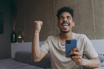 Young african american man wear beige t-shirt sit on grey sofa indoors apartment point index finger...