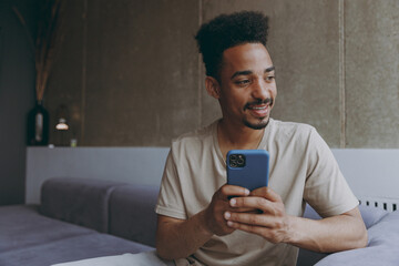 Dreamful pensive young african american man in beige t-shirt sit on grey sofa indoors apartment using mobile cell phone chat online speak with girlfriend looking aside rest on weekends stay at home