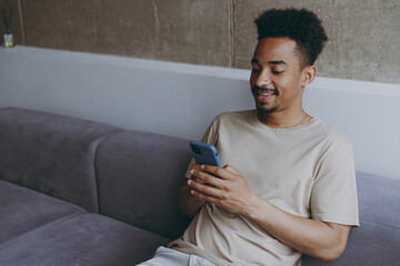 Excited smiling fun happy young african american man wear beige t-shirt sit on grey sofa indoors apartment using mobile cell phone type message sms to friends rest on weekends quarantine stay at home