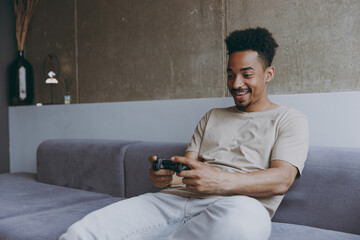 Happy gambling smiling excited overjoyed young african american man in beige t-shirt sit on grey sofa indoors apartment playing pc game with joystick console resting on quarantine weekends stay home.