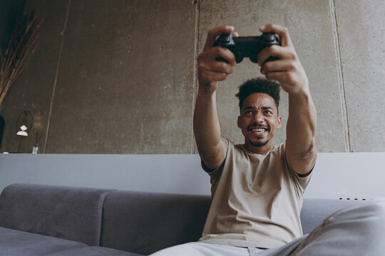 Bottom view epressive addicted young african american man 20s in beige t-shirt sit on grey sofa indoors apartment play pc game with joystick console excitedly rest on quarantine weekends stay home