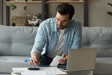 Concentrated young 30s man in eyeglasses calculating expenditures, holding paper bank bills in...