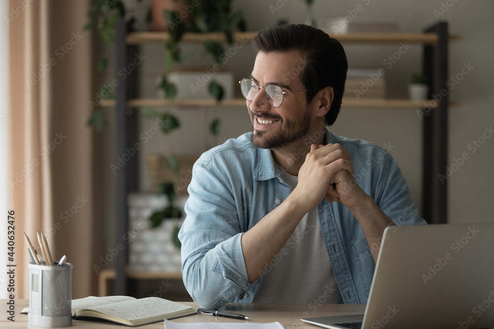 Wall mural smiling positive young man in eyeglasses sitting at table with computer, daydreaming looking in dist