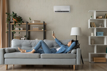 Full length relaxed young happy woman homeowner lying on comfortable sofa turning on air...