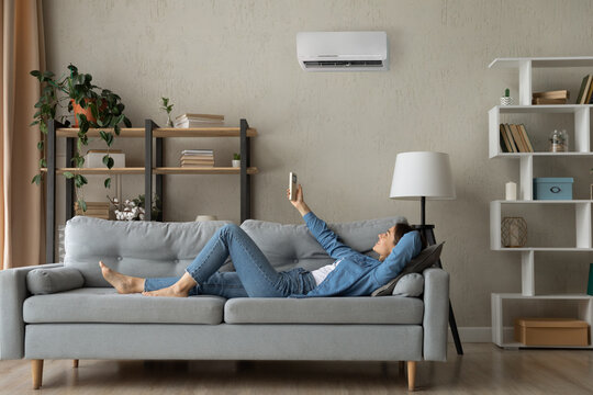 Full length relaxed young happy woman homeowner lying on comfortable sofa turning on air conditioner with remote controller, breathing fresh air, enjoying lazy weekend time, meditating in living room.