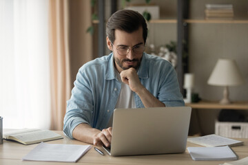 Fototapeta premium Concentrated happy young business man in eyewear looking computer screen, developing online project growth strategy, preparing presentation, web surfing information doing research, working distantly.