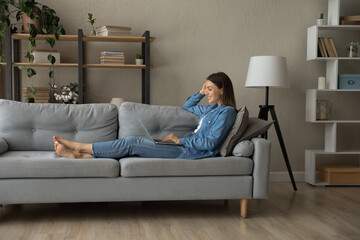 Full length relaxed millennial smiling woman lying on cozy sofa, enjoying using computer software...