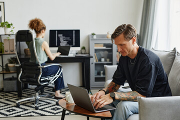 Mature businessman sitting on sofa and working on laptop with his colleague in the background