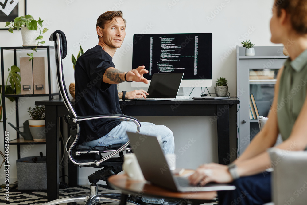 Wall mural programmer discussing new software with his colleague while she sitting on sofa and working on lapto