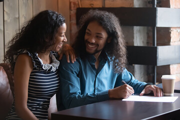 Sincere smiling young african american family couple discussing decision, signing contract with real estate or travel agent, purchasing own dwelling, buying travel tour at meeting in cafe or office.