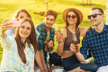 group of young people sitting on a picnic blanket having fun together with pizza and drinks and taking a selfie