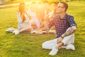 Young man in a plaid shirt, drinking a drink while sitting on the lawn, with a company of friends,...