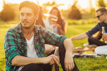 young man, in casual clothes, resting with his friends, sitting on the lawn on a picnic in the park, summer picnic