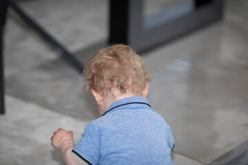 Blonder einjähriger Junge mit vielen Locken sitzt mit dem Rücken zu und spielt , er ist sehr beschäftigt und sieht sehr niedlich aus. Der Boden ist grau, sehr modern.  - obrazy, fototapety, plakaty