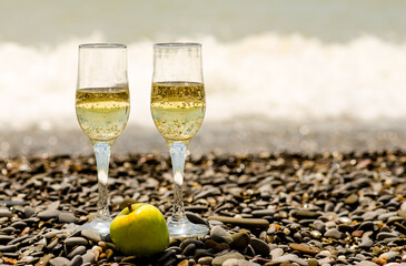 two glasses of champagne on the seashore with a rocky, pebble beach, and a green apple in a clear...