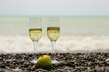 two glasses of champagne on the seashore with a rocky, pebble beach, and a green apple in a clear...