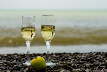 two glasses of champagne on the seashore with a rocky, pebble beach, and a green apple in a clear sunny weather
