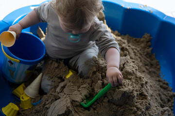 Kleiner Junge mit blonden Locken spielt im blauen Sandkasten. Er bückt sich mit dem Kopf nach unten. Der Junge hat beige Hose und beige Hose an. In der Hand eine Schaufel.