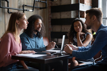 Motivated happy young mixed race employees sitting at table in modern cafe, discussing working issues or online project, developing startup, preparing corporate task, professional teamwork concept.
