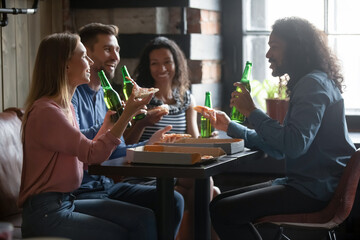 Overjoyed candid millennial multiracial diverse friends drinking beer and eating yummy pizza, enjoying pleasant conversation, communicating on weekend, enjoying leisure hanging out together in cafe.