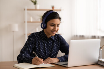 Happy millennial Indian teen student in headphones study online on laptop at home. Smiling smart young mixed race female in earphones look at computer screen make notes working on gadget.