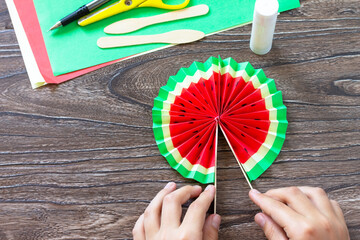 In the hands of a child Paper Fan watermelon on a wooden table. Childrens art project, handmade,...