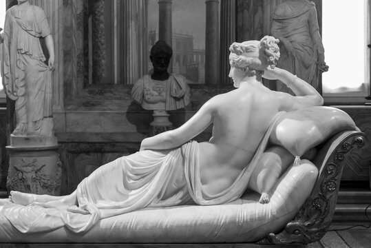 Black and white photo of marble sculpture of a beautiful woman laying down on a divan, viewed from her naked back