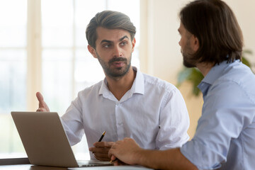 Confident businessman discussing project strategy with business partner, mentor coach training trainee, using laptop, pointing at screen, helping with software, coworkers working together