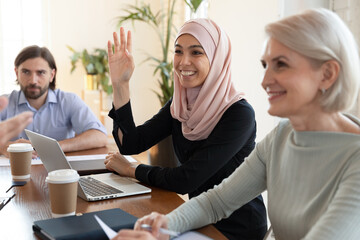 Smiling Asian muslim businesswoman wearing hijab raising hand close up, asking question at meeting...