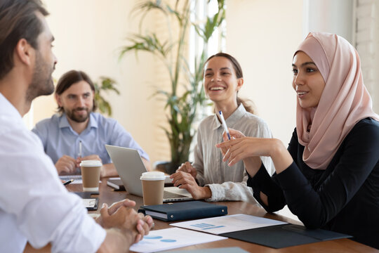 Happy diverse employees, partners discussing startup ideas or project statistics at meeting, smiling Asian muslim businesswoman wearing hijab talking with businessman, having fun, sitting at table