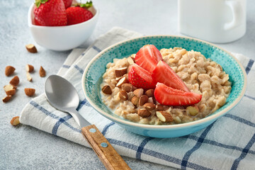 Oatmeal porridge with strawberry slices, nuts almonds and honey in blue bowl on grey table. Healthy eating, dieting, vegetarian food concept. Place for text