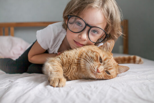 little girl with glasses lies on the bed and hugs a fat ginger cat. High quality photo