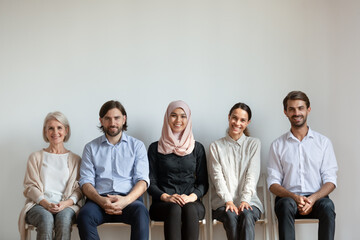 Portrait smiling diverse business people, multiethnic job applicants sitting on chairs in queue, row, waiting for job interview, looking at camera, staff, human resources and employment concept